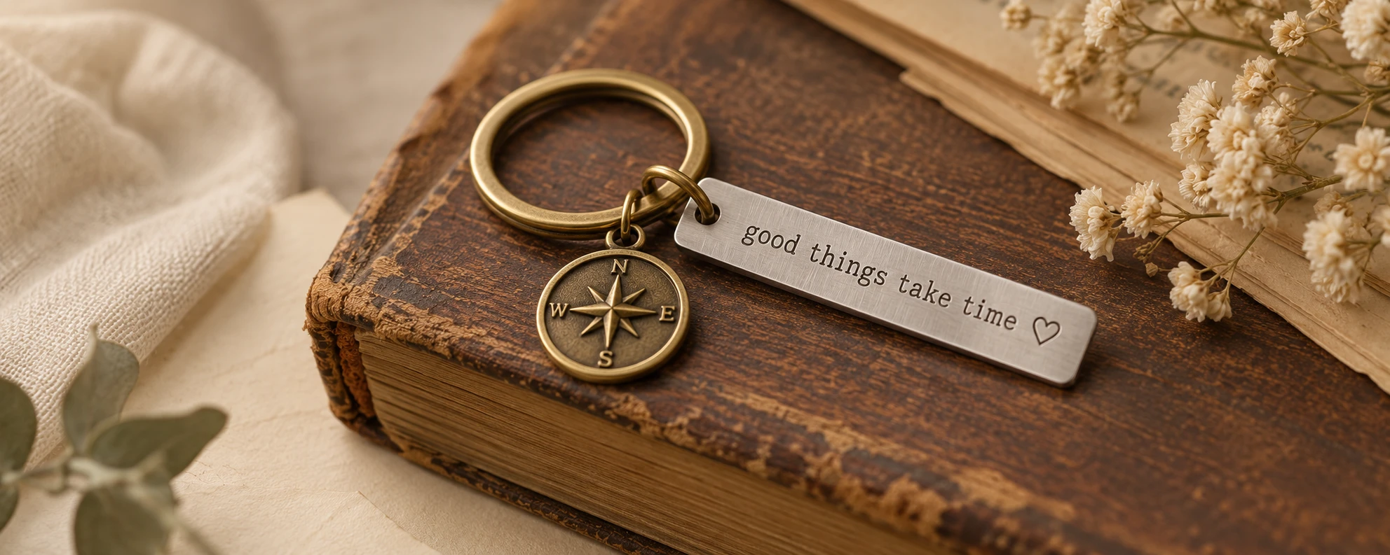 A silver message keychain and brass compass charm on a weathered book with cream linen and dried flowers